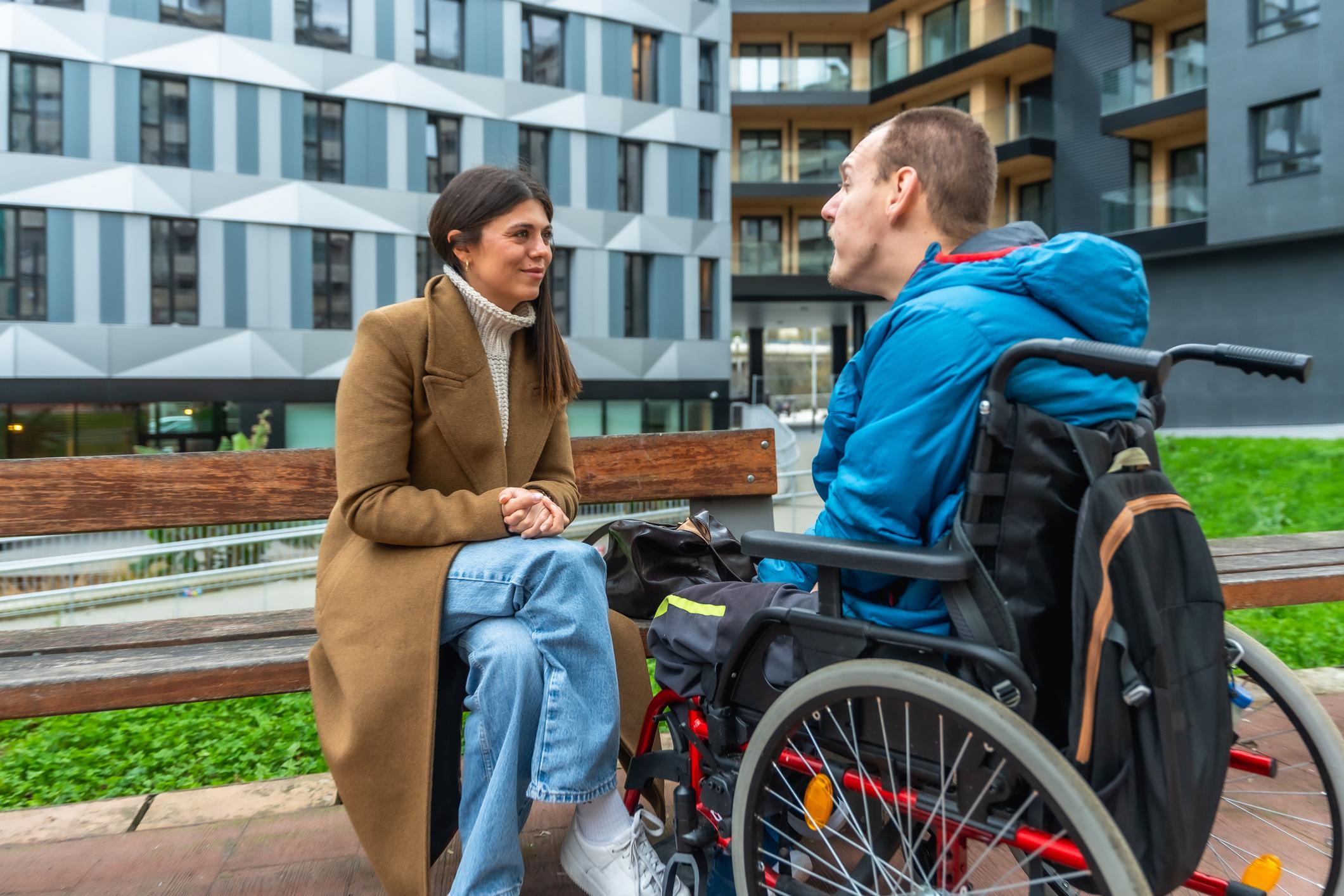 Young adult woman sitting on a park bench talking with a mature adult man using a wheelchair, showing support, care, and inclusivity in an urban environment