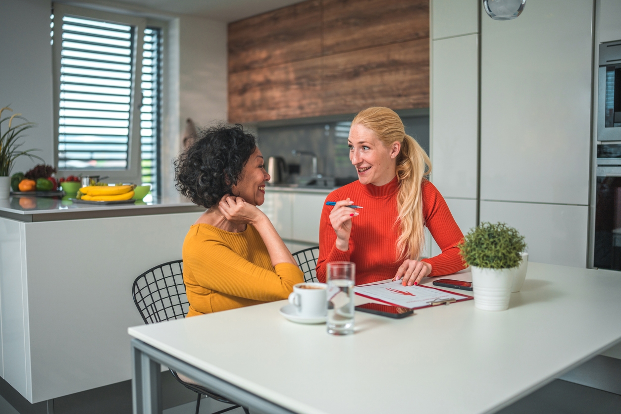 Waist up image of a Hispanic mature woman sitting at a table with a professional female nutritionist, talking about a healthier lifestyle. Modern kitchen in the background.