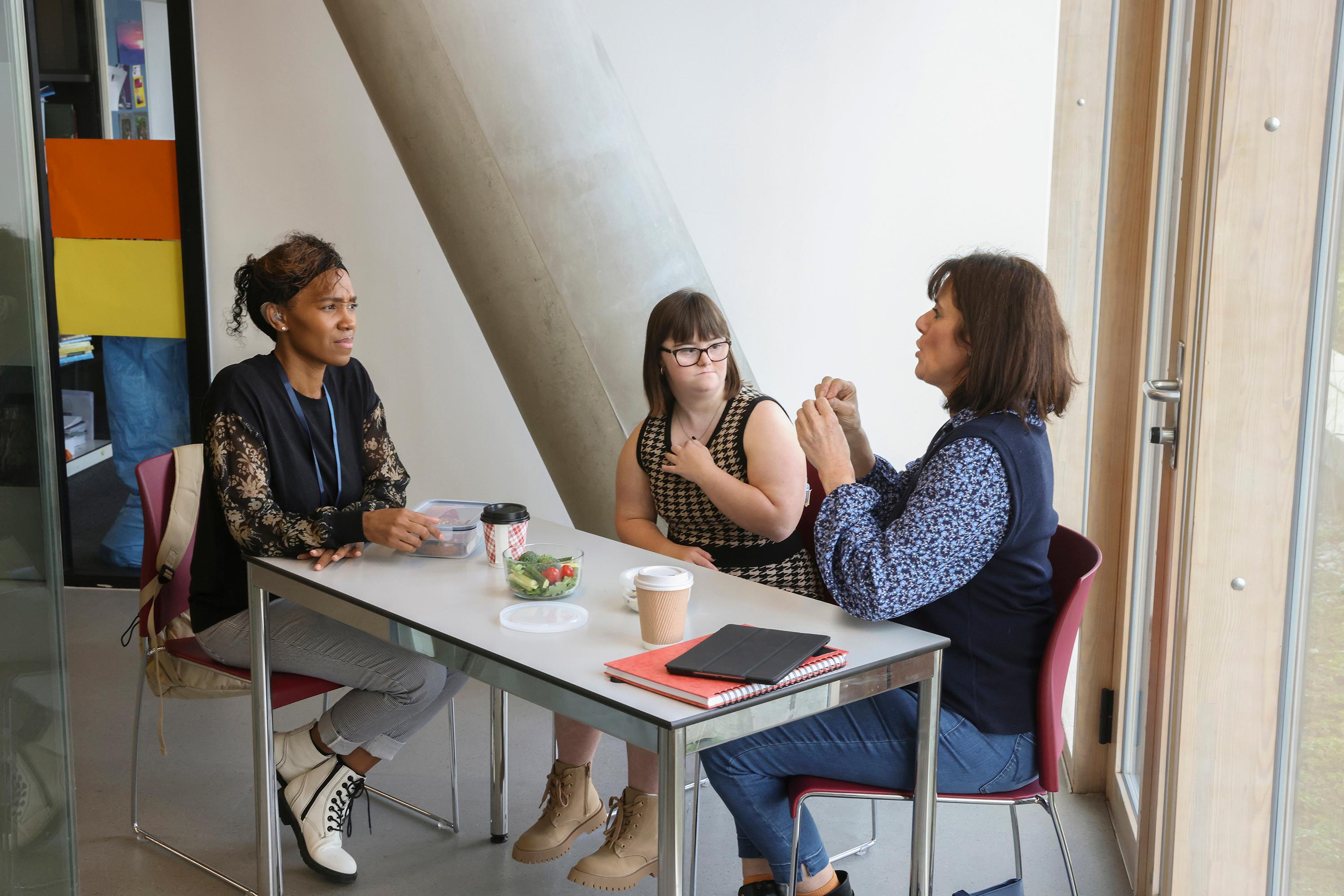 Women, including woman with Down Syndrome, using sign language during lunch at table in office
